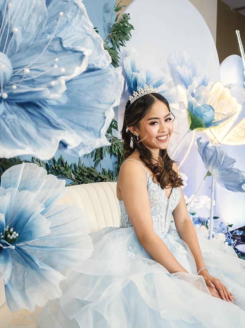 A debutante in a pale blue gown on a white sofa inside a wall of oversized blue flowers