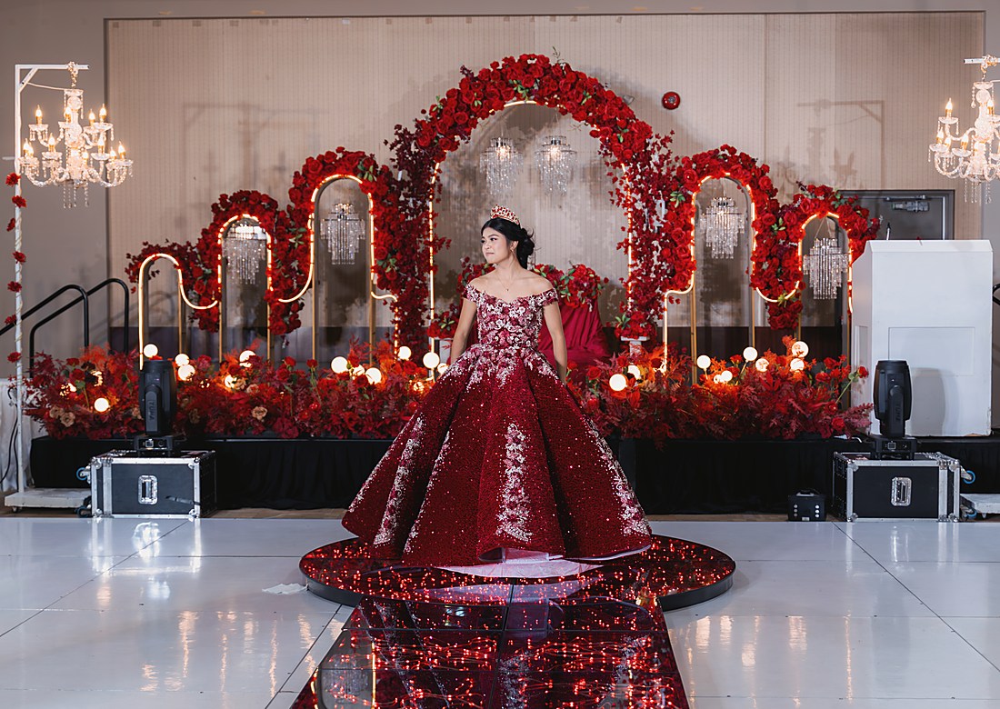 A debutante in a red ball gown crossing a mirrored floor beneath a red floral arch