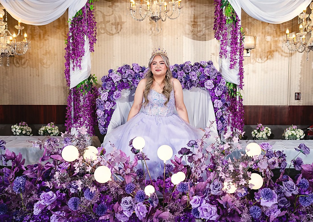 A debutante seated on a throne inside a wall of purple and white flowers