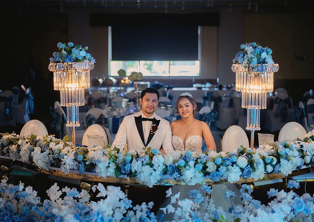 A couple at their head table behind a bank of white and blue flowers