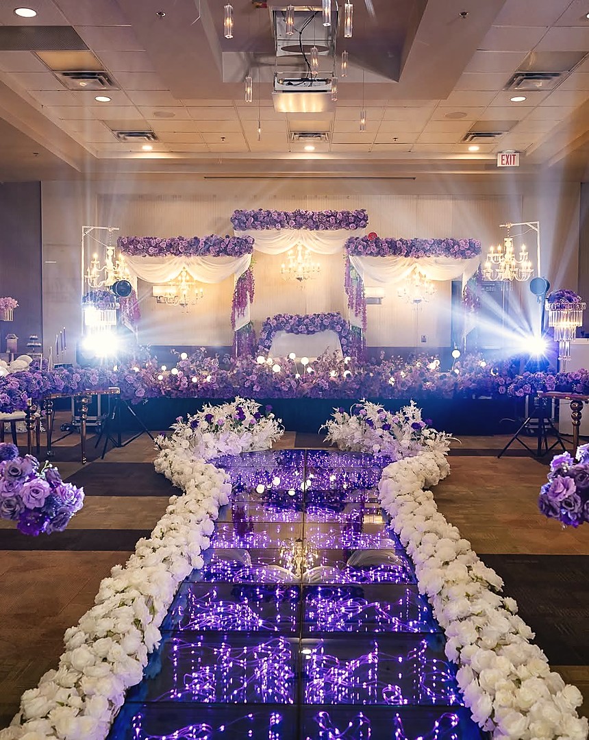 A ballroom aisle of lit mirror and white flowers running to a purple floral stage