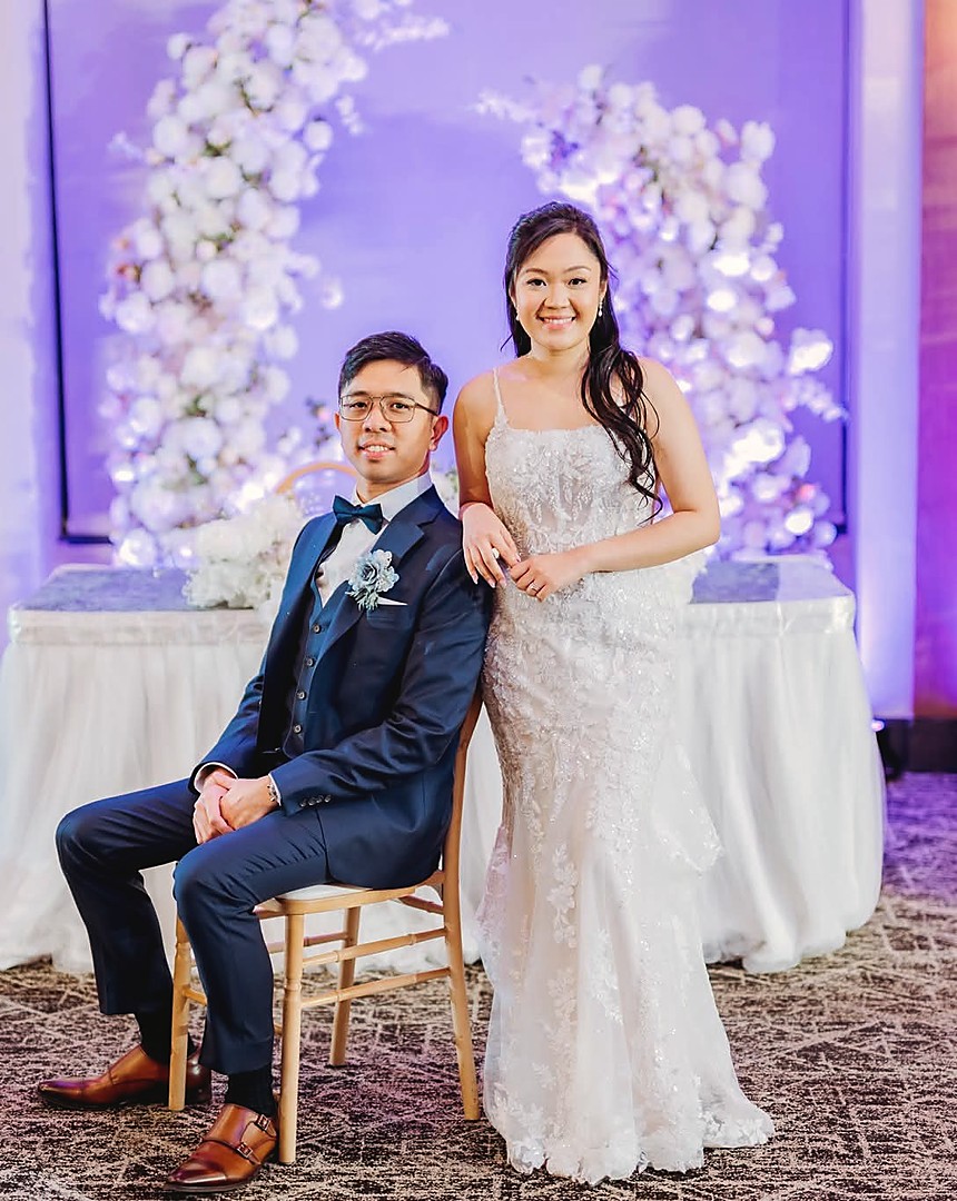 A couple at their head table in front of a wall of white orchids
